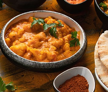 Curry de Chou-fleur Indien Savoureux et Épicé Bol de curry de chou-fleur crémeux et orange, garni de persil frais, accompagné de pain plat et d'épices rouges en poudre.